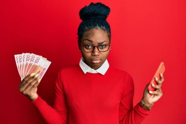 Genç Afro-Amerikalı kadın elinde Norveç Kronu ile akıllı telefon kullanıyor şüpheci ve sinirli, sorun yüzünden üzgün. negatif kişi. 