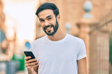 Genç İspanyol adam şehirde akıllı telefon kullanarak mutlu gülümsüyor..