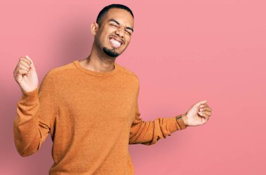 Genç Afro-Amerikan erkeği gündelik kıyafetler giymiş mutlu ve neşeli dans ediyor, gülümsüyor rahat ve kendinden emin bir şekilde müzik dinliyor. 