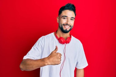 Sakallı, müzik dinleyen, kulaklık takan, mutlu bir el hareketi yapan genç bir adam. Kameraya bakıp başarıyı gösteren ifade. 