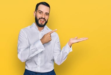 Sakallı, iş gömleği giyen genç adam kameraya hayret ediyor ve gülümsüyor.. 