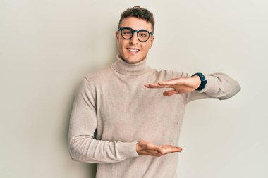 Balıkçı yaka kazak giyen İspanyol genç bir adam. Elleriyle büyük ve büyük bir işaret, ölçü sembolü gösteriyor. Kameraya bakıp gülümsüyor. ölçüm kavramı. 