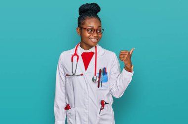 Genç Afro-Amerikan kadın doktor üniforması ve steteskop takmış gülümseyen, gülümseyen ve baş parmağıyla yüzünü gösteren.. 