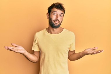 Sarı arka planda sıradan sarı tişörtler giyen sakallı yakışıklı ve elleri ve kolları havada, kafası karışmış bir adam. şüphe kavramı. 