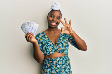 Kıllı türban takan genç Afrikalı bir kadın elinde 20 İsveç kronu banknotuyla tamam işareti yapıyor, gülümseyerek mükemmel bir sembol oluşturuyor. 