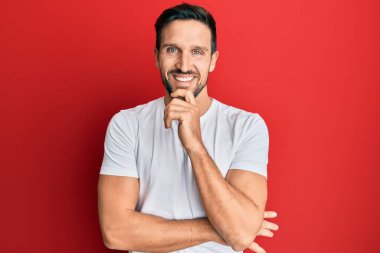 Gündelik beyaz tişört giyen genç yakışıklı adam kameraya kollarını kavuşturmuş ve çenesi üzerinde kendinden emin bir şekilde bakıyor. Olumlu düşünmek. 