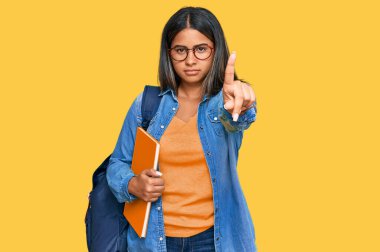 Öğrenci sırt çantası takan, elinde kitaplarla elini havaya kaldırmış ve kızgın bir ifade sergileyen genç Latin bir kız. 