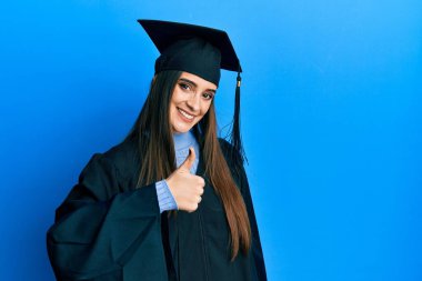 Mezuniyet şapkası ve tören cübbesi takan güzel esmer bir kadın mutlu bir el hareketi yapıyor. Kameraya bakıp başarıyı gösteren ifade. 