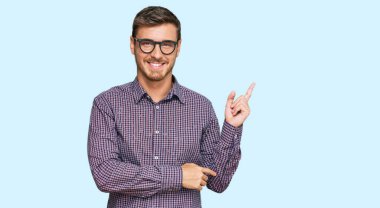 Yakışıklı beyaz adam, günlük kıyafetler ve gözlük takıyor. Yüzünde kocaman bir gülümseme var. Eli ve parmağı kameraya bakıyor.. 