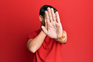 Yakışıklı, beyaz bir adam, günlük kırmızı tişört giyiyor, elleriyle gözlerini kapatıyor ve üzgün ve korku ifadeleriyle jest yapıyor. utangaç ve olumsuz kavram. 