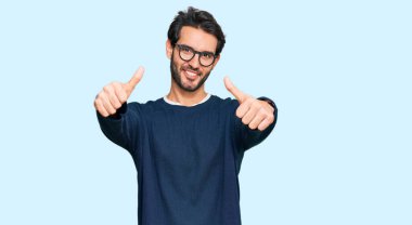 Gündelik kıyafetler giyen ve gözlük takan genç İspanyol adam elinle olumlu jest yapıyor, gülümsüyor ve başarı için mutlu. kazanan jest. 