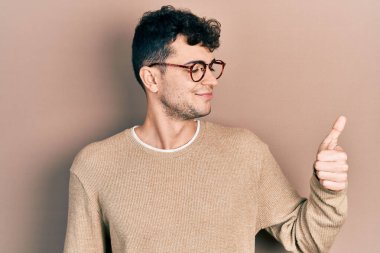 Gündelik kıyafetler ve gözlük takan genç İspanyol adam gururlu görünüyor, gülümsüyor yan tarafa baş parmak hareketi yapıyor. 