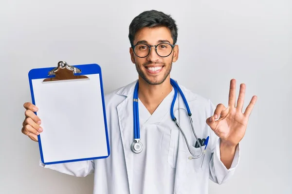 Jovem Homem Bonito Vestindo Médico Estetoscópio Segurando Prancheta Fazendo Sinal — Fotografia de Stock