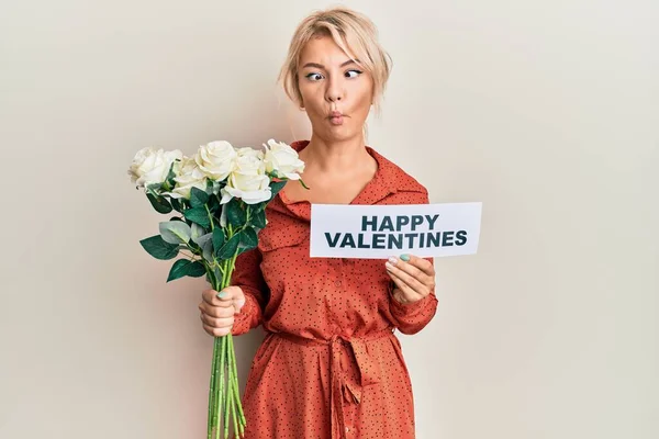Young blonde girl holding flowers and paper with happy valentines message making fish face with mouth and squinting eyes, crazy and comical.