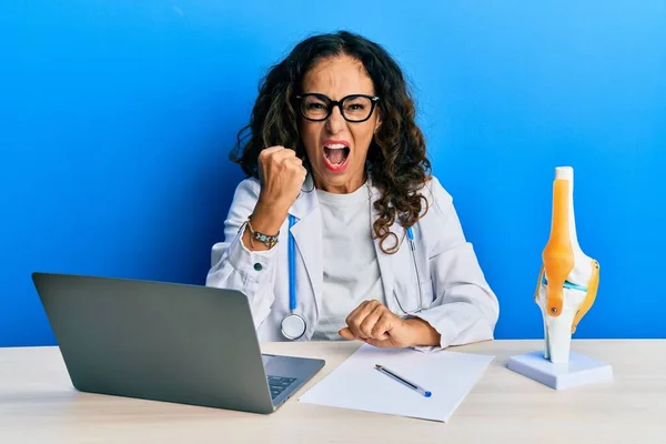 Beautiful middle age woman doctor at orthopedic clinic angry and mad raising fist frustrated and furious while shouting with anger. rage and aggressive concept.