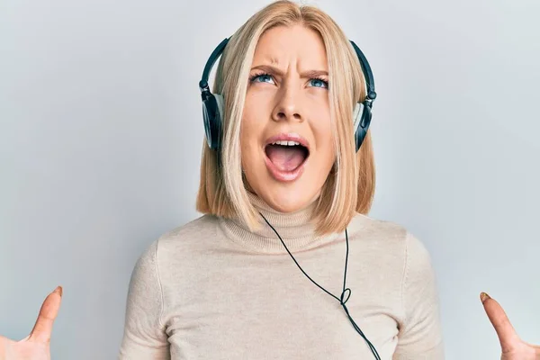 ヘッドフォンを使用して音楽を聴いている若いブロンドの女性は 狂気と狂気の叫びと積極的な表現と腕を上げて叫んでいます フラストレーションコンセプト — ストック写真