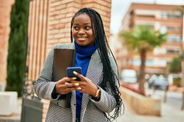 Genç Afrikalı Amerikalı iş kadını şehirde akıllı telefon kullanarak mutlu bir şekilde gülümsüyor..