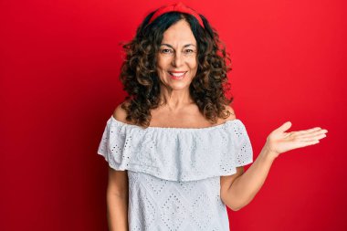 Orta yaşlı İspanyol kadın gündelik kıyafetler giyiyor neşeli bir şekilde gülümsüyor ve avuç içiyle kameraya bakıyor.. 