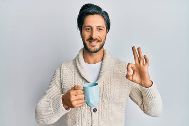 Genç İspanyol adam bir fincan kahve içiyor parmaklarıyla imza atıyor, gülümsüyor ve mükemmel bir sembol oluşturuyor. 