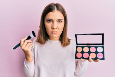 Makyaj fırçası tutan genç ve güzel bir kadın. Yüzü kızardı, şüpheci ve gergin. Problemden dolayı surat asıyor. negatif kişi. 