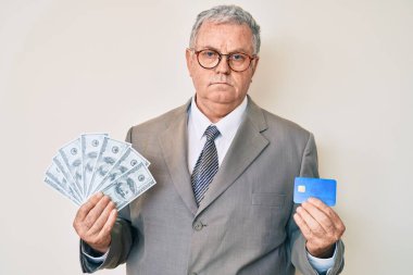 Üstün gri saçlı, takım elbiseli, kredi kartı takan ve yüzünde ciddi bir ifadeyle dolar taşıyan bir adam. Basit ve doğal bir şekilde kameraya bakıyorsun.. 