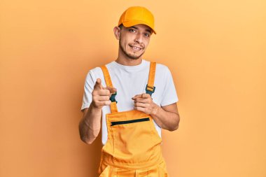 Tamirci üniforması giyen İspanyol genç adam mutlu ve komik yüzüyle kamerayı işaret ediyor. iyi enerji ve titreşimler. 