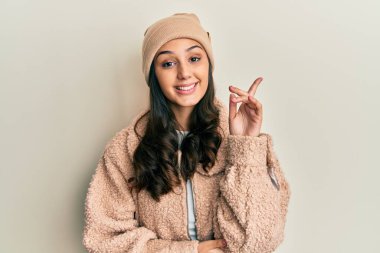 Yün kazak giyen ve yüzünde kocaman bir gülümseme olan genç İspanyol kadın kameraya el ve parmağıyla bakıyor.. 