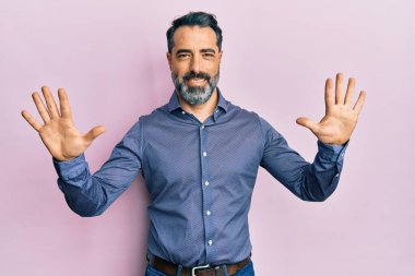 Sakallı, gri saçlı, iş kıyafetleri giyen orta yaşlı bir adam on numaralı parmaklarıyla işaret ederken kendinden emin ve mutlu bir şekilde gülümsüyor.. 