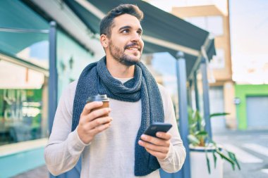 Genç İspanyol adam mutlu bir şekilde gülümsüyor. Akıllı telefon kullanıyor ve şehirde kahve içiyor..