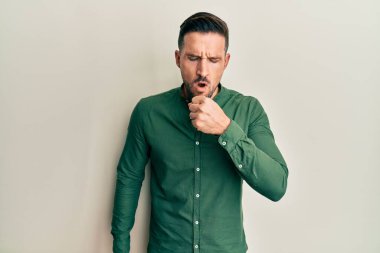 Sakallı, sıradan kıyafetler giyen, kendini kötü hisseden ve soğuk algınlığı veya bronşitin belirtisi olarak öksüren yakışıklı bir adam. sağlık hizmeti kavramı. 