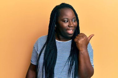 Saçları örgülü genç siyahi bir kadın başparmağını kaldırmış, ağzı açık gülümsüyor. 