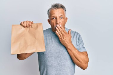 Orta yaşlı, kır saçlı, paket kağıdı tutan, ağzı elle kaplayan, şok olmuş ve hata yapmaktan korkan bir adam. şaşırmış ifade 