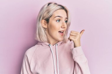 Sıradan kıyafetler giyen genç sarışın kız baş parmağını yana kaldırarak mutlu bir şekilde ağzını açarak gülümsüyor. 