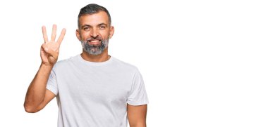 Orta yaşlı, beyaz tişört giyen yakışıklı bir adam üç numarayı gösterip işaret ederken kendinden emin ve mutlu bir şekilde gülümsüyor.. 
