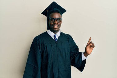 Yakışıklı siyah adam mezuniyet şapkası ve tören cübbesi takıyor. Yüzünde kocaman bir gülümseme var. Parmağıyla kameraya bakıyor.. 