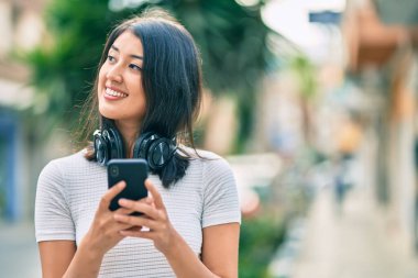 Genç İspanyol kadın şehirde akıllı telefon ve kulaklık kullanarak mutlu bir şekilde gülümsüyor..
