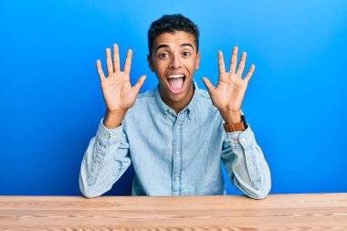 Genç, yakışıklı, Afro-Amerikalı, gündelik giysiler giymiş. Masada oturuyor ve 10 numarayı işaret ediyor. Kendinden emin ve mutlu bir şekilde gülümsüyor.. 