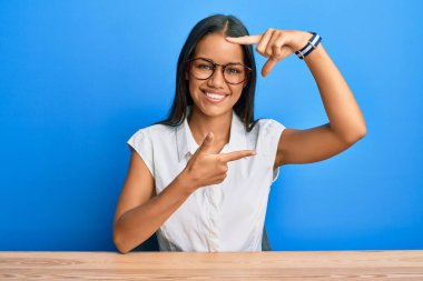 Güzel İspanyol kadın, gündelik giysiler giyip masaya oturmuş gülümsüyor ve elleri ve parmakları mutlu yüzlü bir çerçeve yapıyor. yaratıcılık ve fotoğrafçılık kavramı. 