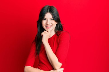 Genç, beyaz, sıradan kırmızı tişörtlü bir kız kameraya kollarını kavuşturup, çenesine dokunarak gülümsüyor. Olumlu düşünmek. 