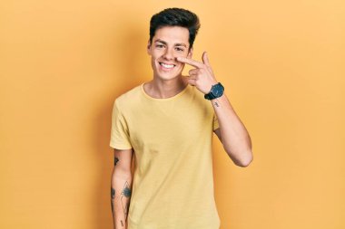Sarı günlük tişörtler giyen genç İspanyol adam el ve yüz parmaklarını ve burnunu göstererek neşeli bir şekilde gülümsüyor. güzellik konsepti 