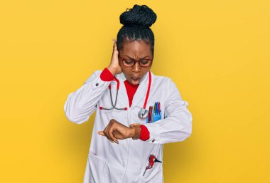Genç Afrikalı Amerikalı kadın doktor üniforması ve steteskop giyiyor. Saate bakıyor. Endişeli, geç kalmaktan korkuyor. 