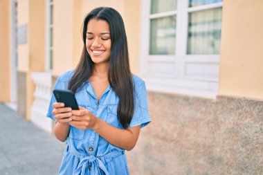 Genç Latin kız şehirde akıllı telefon kullanarak mutlu bir şekilde gülümsüyor..