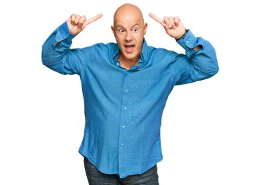Orta yaşlı, kel, sıradan kıyafetler giyen, şaşkın, şaşkın, parmaklarıyla işaret eden ve kollarını kaldıran bir adam.. 