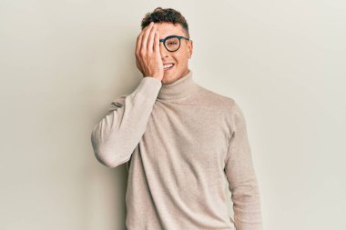 Balıkçı yaka kazak giyen İspanyol genç bir adam bir gözünü eliyle kapatıyor, yüzünde kendine güvenen bir gülümseme ve şaşırtıcı bir duygu var.. 