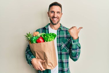 Elinde poşet sakallı yakışıklı bir adam. Elinde alışveriş poşeti, arkasında da el ve baş parmakları var. Kendinden emin gülümsüyor. 