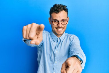 Gündelik kıyafetler giyen ve gözlüklü genç İspanyol adam seni ve kamerayı parmaklarıyla göstererek mutlu ve mutlu gülümsüyor. 