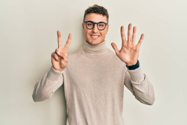 Balıkçı yaka kazak giyen İspanyol genç adam yedinci parmaklarını gösterip gösterirken kendinden emin ve mutlu bir şekilde gülümsüyor.. 