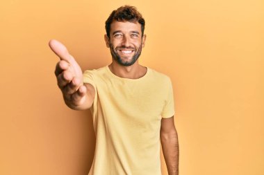 Sarı arka planda sıradan sarı tişörtler giyen sakallı yakışıklı bir adam selamlama ve hoş geldin demek için gülümseyerek el sıkışır. başarılı iş. 