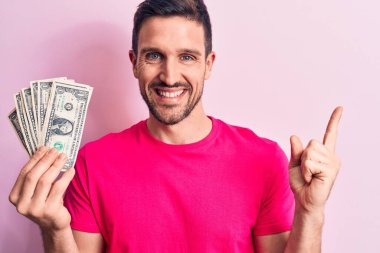 Soyutlanmış pembe arka planda bir deste banknot tutan yakışıklı genç adam mutlu bir şekilde elini ve parmağını kenarı işaret ediyor.