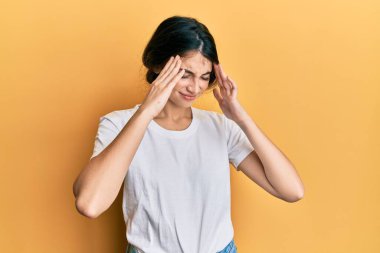 Beyaz gündelik tişörtler giyen, baş ağrısı çeken, çaresiz ve stresli genç beyaz bir kadın. Çünkü acı ve migren ağrısı var. Eller başının üstünde.. 